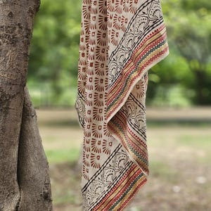 May include: A beige textile with a repeating brown floral pattern and a decorative border. The border features black, red, yellow, and green stripes. The fabric is draped over a tree branch against a blurred green background.
