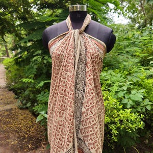 May include: A beige and red patterned sarong is draped on a mannequin. The sarong features a repeating geometric design in red and beige, with black and red borders. The fabric is tied at the neck, showcasing the full length of the garment.