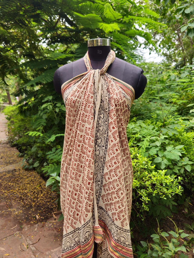 May include: A beige and red patterned sarong is draped on a mannequin. The sarong features a repeating geometric design in red and beige, with black and red borders. The fabric is tied at the neck, showcasing the full length of the garment.