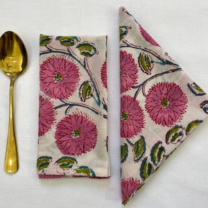 Op de afbeelding: Een gouden lepel ligt naast twee gevouwen witte stoffen servetten. De servetten hebben een patroon van grote roze bloemen met groene bladeren en blauwgroene stelen. De servetten zijn waarschijnlijk voor het diner.