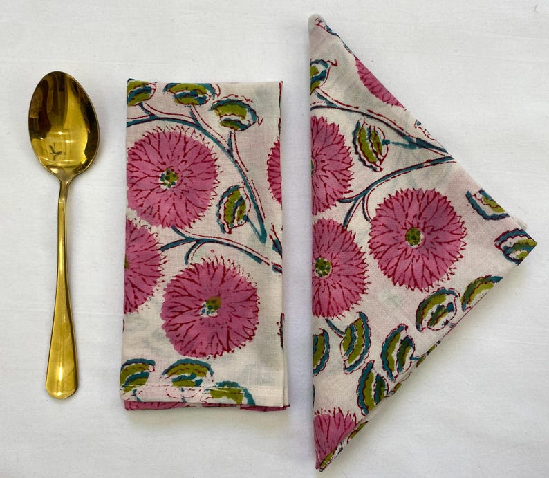 Op de afbeelding: Een gouden lepel ligt naast twee gevouwen witte stoffen servetten. De servetten hebben een patroon van grote roze bloemen met groene bladeren en blauwgroene stelen. De servetten zijn waarschijnlijk voor het diner.