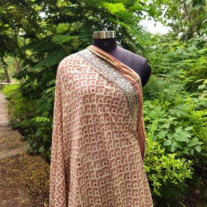 May include: A beige shawl with a repeating geometric pattern in reddish-brown. The shawl is draped over a black mannequin. The border features a black, red, and yellow design. The background is a lush green outdoor setting.