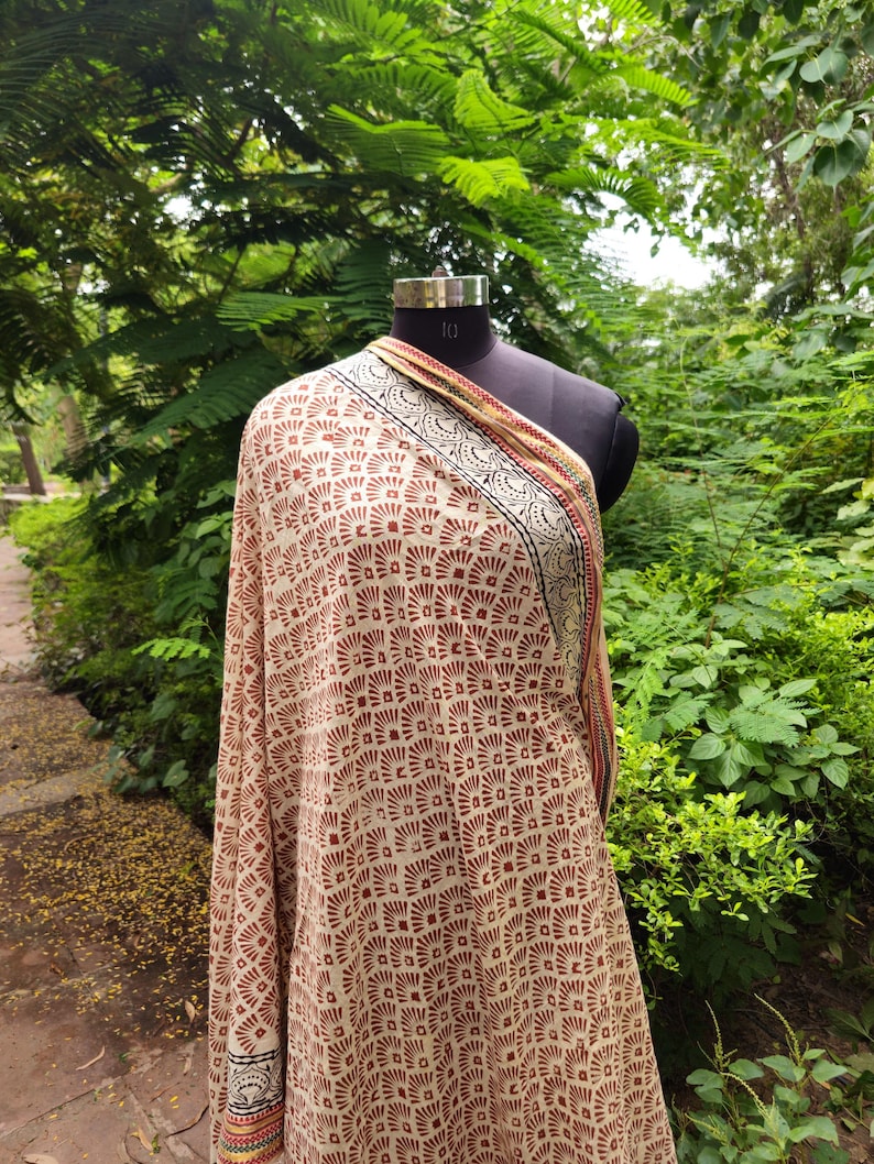 May include: A beige shawl with a repeating geometric pattern in reddish-brown. The shawl is draped over a black mannequin. The border features a black, red, and yellow design. The background is a lush green outdoor setting.