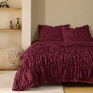 May include: A deep burgundy comforter set featuring a heavily ruched, textured surface with horizontal gathered detailing. The set includes two matching rectangular pillow shams and a large comforter, all finished with a row of small buttons.
