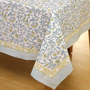 Könnte beinhalten: Weißes Tischtuch mit floralem Muster in Blau- und Gelbtönen. Das Design zeigt stilisierte Blumen und Blätter. Der Rand hat ein kariertes Muster in Hellblau und eine dekorative gelbe Bordüre. Das Tischtuch ist über einen Holztisch drapiert.