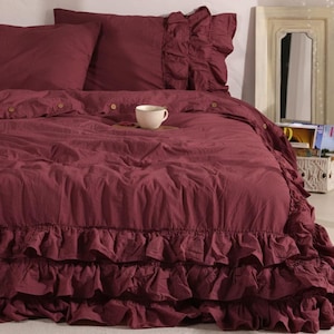 May include: A burgundy bed set with a ruffled duvet cover and pillowcases. The bedding features wooden button details and a white teacup on a small wooden tray. The bed is made up with a layered look, creating a cozy and inviting aesthetic.