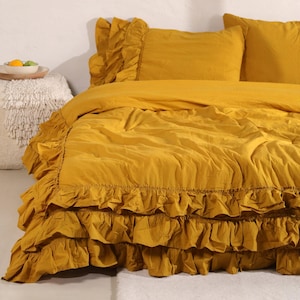 May include: Mustard yellow bedding set featuring a ruffled duvet cover and pillow shams. The set includes a duvet cover and two pillow shams. The bedding is made of a soft, textured fabric. The bed is against a white wall.