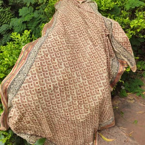 May include: A beige textile with a repeating brown fan-shaped pattern. The edges feature a decorative border with black, red, and yellow accents. The fabric is draped over a green leafy background.