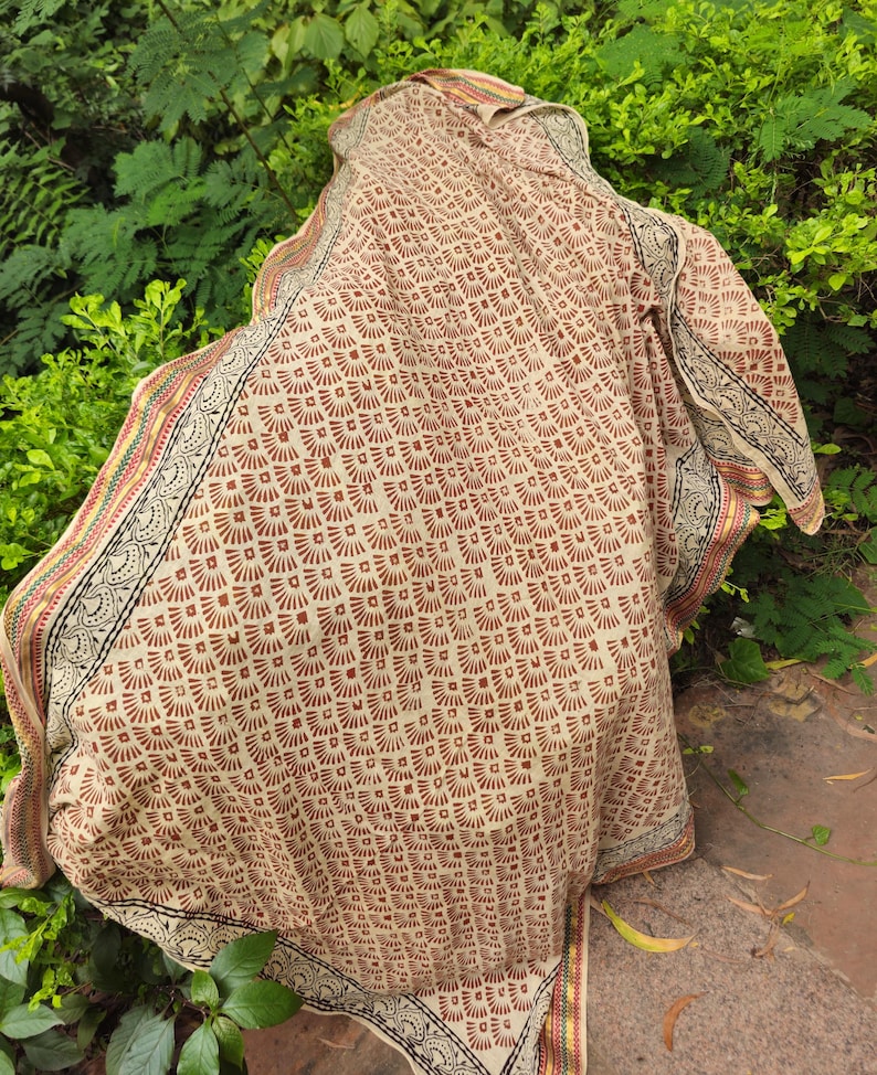 May include: A beige textile with a repeating brown fan-shaped pattern. The edges feature a decorative border with black, red, and yellow accents. The fabric is draped over a green leafy background.