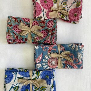 May include: Four sets of floral print cloth napkins tied with beige ribbons. The napkins feature various designs in pink, blue, and white, with floral and botanical patterns. The napkins are neatly stacked, suggesting a home decor or dining theme.