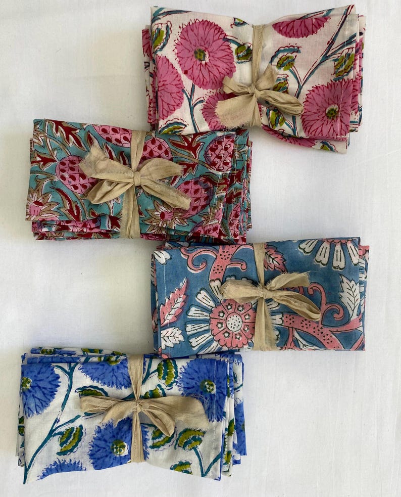 May include: Four sets of floral print cloth napkins tied with beige ribbons. The napkins feature various designs in pink, blue, and white, with floral and botanical patterns. The napkins are neatly stacked, suggesting a home decor or dining theme.