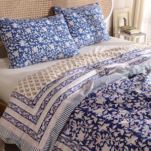 May include: A blue and white floral patterned bedding set. The set includes two square pillowcases and a quilt. The quilt features a mix of floral and striped patterns in blue, white, and beige. The bedding is arranged on a bed with a woven headboard.