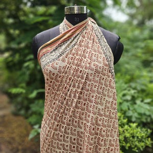 May include: A beige and brown patterned sarong is draped over a black mannequin. The sarong features a repeating geometric design in shades of brown and beige. The top edge has a decorative border with black, red, and gold accents.