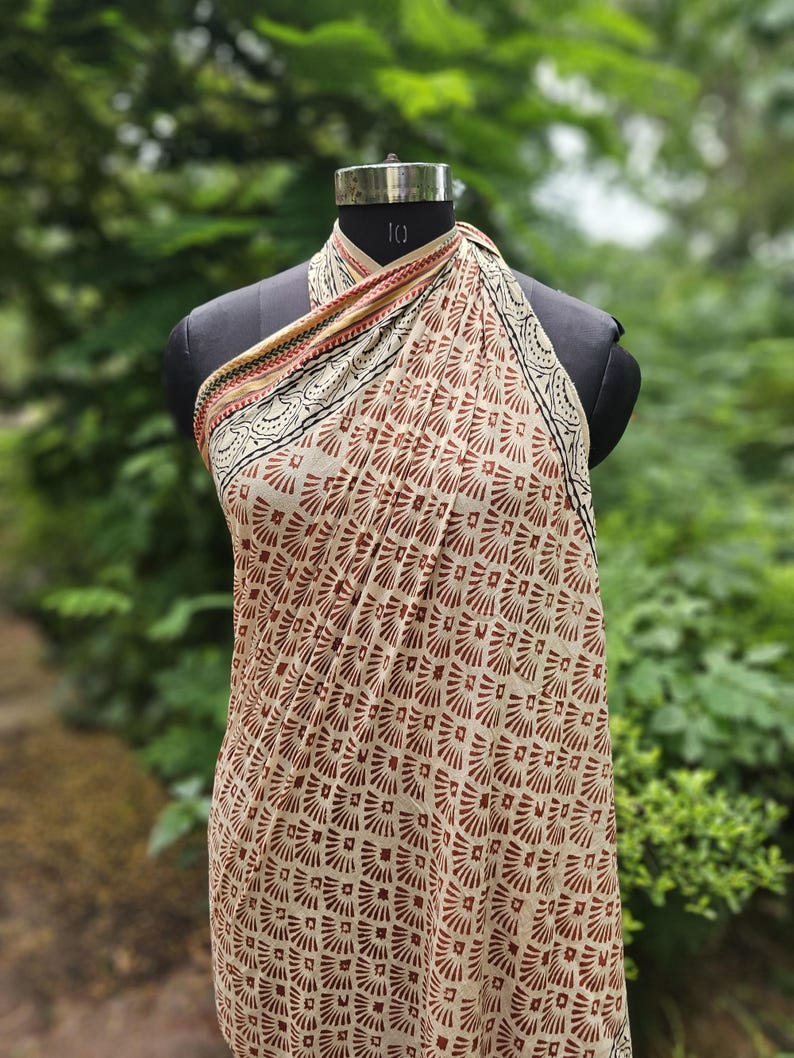 May include: A beige and brown patterned sarong is draped over a black mannequin. The sarong features a repeating geometric design in shades of brown and beige. The top edge has a decorative border with black, red, and gold accents.
