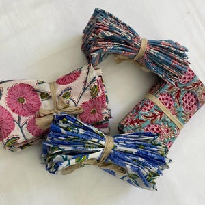 May include: Bundles of folded cloth napkins with floral and fruit patterns. The napkins are tied with twine. The patterns include pink flowers, blue and white flowers, and red pineapples on a teal background.
