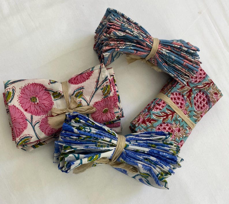 May include: Bundles of folded cloth napkins with floral and fruit patterns. The napkins are tied with twine. The patterns include pink flowers, blue and white flowers, and red pineapples on a teal background.