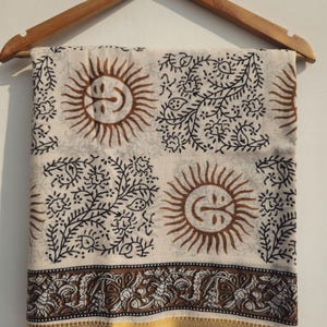 Puede incluir: Tela color crema con un patrón repetido de soles marrones y diseños florales negros. Un borde marrón y dorado recorre el borde inferior. La tela está colgada de una percha de madera.