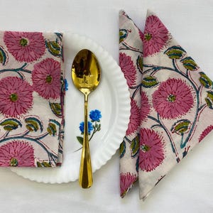 Op de afbeelding: Een gevouwen roze bloemen servet ligt op een wit bord met een gouden lepel. Twee extra servetten met hetzelfde roze bloemenpatroon zijn in een driehoek gevouwen. Het bloemenpatroon bevat roze bloemen, groene bladeren en blauwe ranken.
