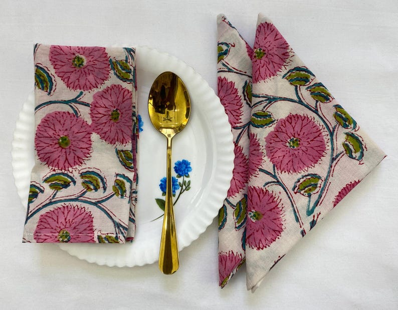 Op de afbeelding: Een gevouwen roze bloemen servet ligt op een wit bord met een gouden lepel. Twee extra servetten met hetzelfde roze bloemenpatroon zijn in een driehoek gevouwen. Het bloemenpatroon bevat roze bloemen, groene bladeren en blauwe ranken.