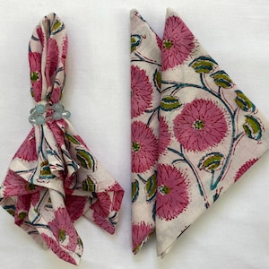 Op de afbeelding: Twee gevouwen stoffen servetten met een bloemenpatroon in roze, groen en blauw op een cr&egrave;mekleurige achtergrond. E&eacute;n servet wordt vastgehouden door een lichtblauwe kralen servetring. De servetten zijn waarschijnlijk voor een tafel.