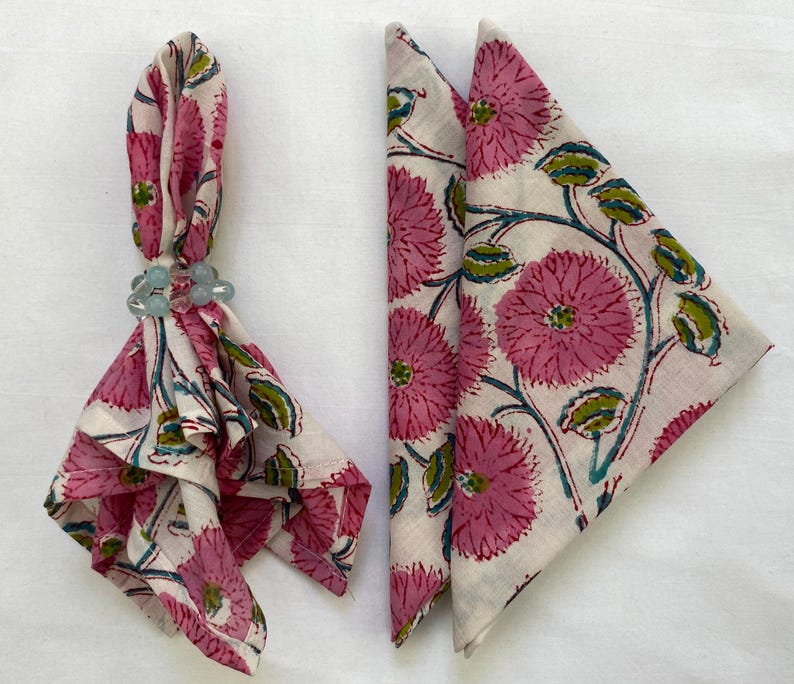 Op de afbeelding: Twee gevouwen stoffen servetten met een bloemenpatroon in roze, groen en blauw op een cr&egrave;mekleurige achtergrond. E&eacute;n servet wordt vastgehouden door een lichtblauwe kralen servetring. De servetten zijn waarschijnlijk voor een tafel.