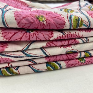Op de afbeelding: Een stapel gevouwen stof met een bloemenpatroon. Het ontwerp toont grote roze bloemen met rode contouren, turkooizen stelen en groene accenten op een witte achtergrond. De stof is gelaagd en toont het herhalende patroon.