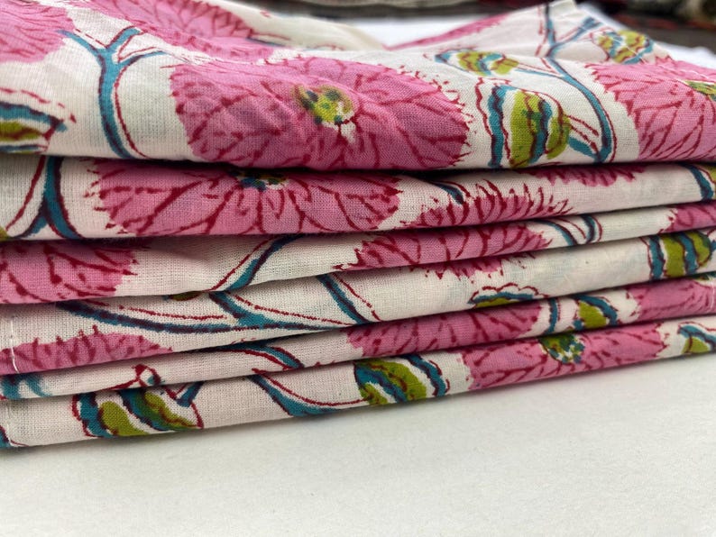 Op de afbeelding: Een stapel gevouwen stof met een bloemenpatroon. Het ontwerp toont grote roze bloemen met rode contouren, turkooizen stelen en groene accenten op een witte achtergrond. De stof is gelaagd en toont het herhalende patroon.