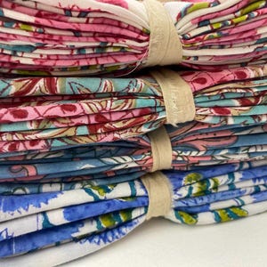 May include: A stack of folded fabric napkins tied with beige ribbon. The napkins feature floral patterns in pink, red, blue, and white. The stack includes a variety of colors and designs, suitable for home decor or special events.