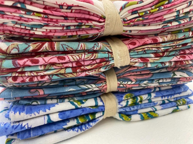 May include: A stack of folded fabric napkins tied with beige ribbon. The napkins feature floral patterns in pink, red, blue, and white. The stack includes a variety of colors and designs, suitable for home decor or special events.