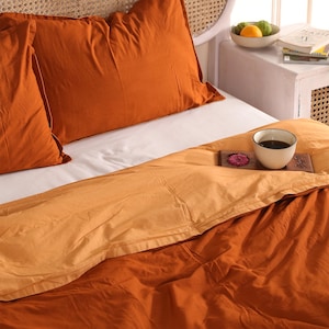 May include: A bed with orange bedding, including two pillowcases and a duvet cover. A white sheet is visible. A cup of coffee and a decorative item sit on a wooden tray on the bed. A side table with fruit and books is in the background.