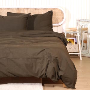 Könnte beinhalten: Ein Bett mit einer dunkelbraunen Bettdecke und passenden Kissenbezügen. Das Kopfteil besteht aus geflochtenem Rattan. Ein kleiner weißer Nachttisch mit Büchern und einem dekorativen Spiegel ist zu sehen.