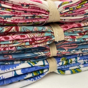 May include: A stack of folded floral patterned fabric napkins in pink, blue, and white, bound with beige bands. The napkins feature a variety of floral designs, creating a colorful and decorative display.