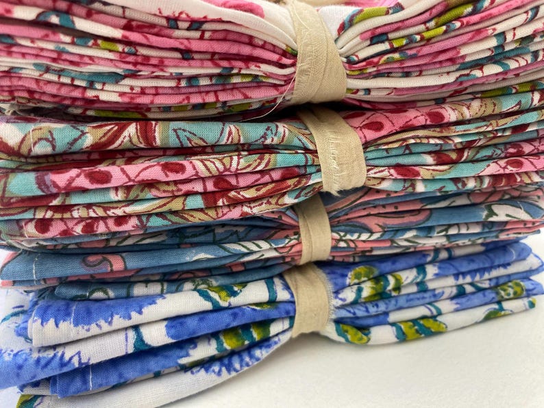 May include: A stack of folded floral patterned fabric napkins in pink, blue, and white, bound with beige bands. The napkins feature a variety of floral designs, creating a colorful and decorative display.