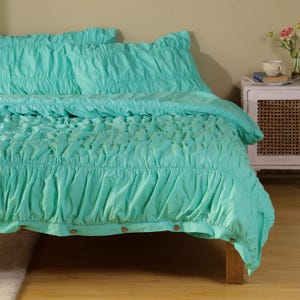 May include: A turquoise bedspread and matching pillowcases with a gathered, textured design. The bedspread is draped over a wooden bed frame. The bedding set is a solid color and has a soft, inviting appearance.