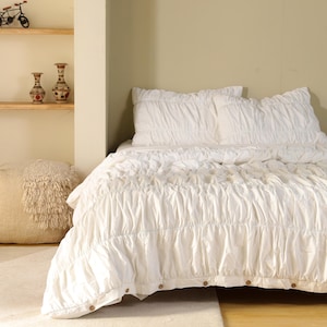 May include: A white bedspread and pillow set with a textured, gathered design. The bedspread has a ruffled edge and is secured with wooden buttons. The bedding is on a bed in a room with a neutral color palette.