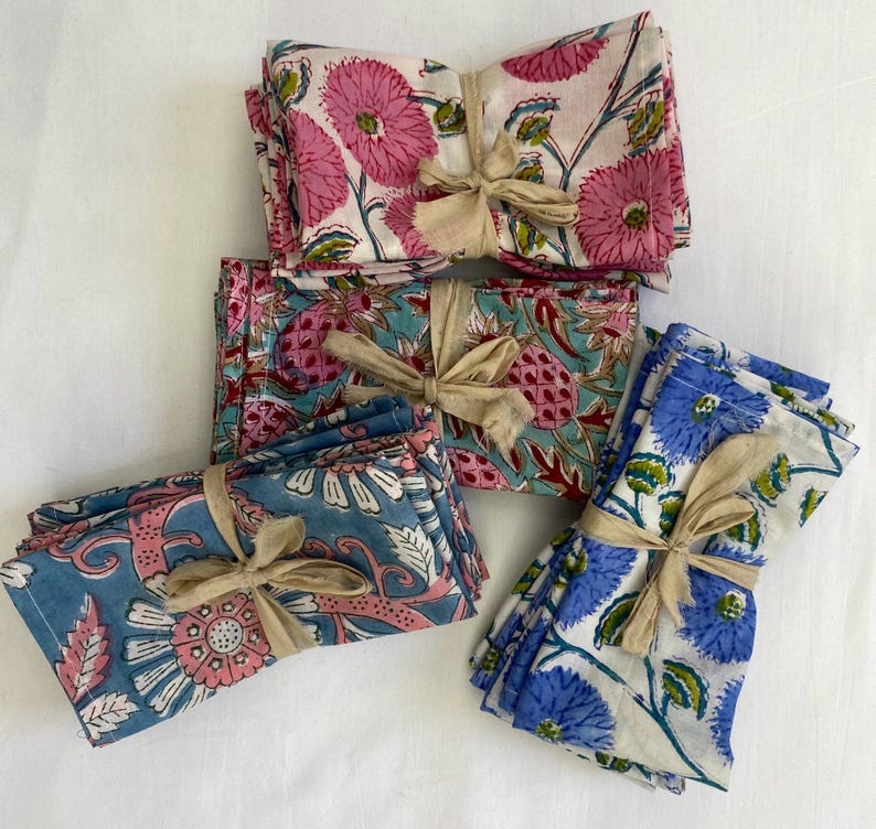 May include: Bundled sets of floral print cloth napkins tied with beige ribbons. The napkins feature pink, blue, and teal floral patterns on a white background. The napkins are made of a soft, lightweight fabric.