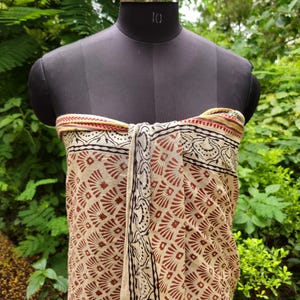 May include: A beige and brown patterned sarong is draped over a black mannequin. The sarong features a repeating geometric design in brown and black, with a decorative border. The background is a lush green foliage.