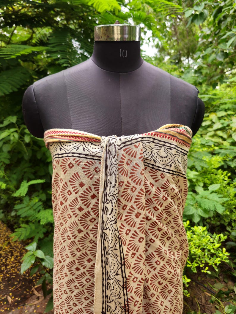 May include: A beige and brown patterned sarong is draped over a black mannequin. The sarong features a repeating geometric design in brown and black, with a decorative border. The background is a lush green foliage.