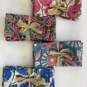 May include: Bundled sets of floral print napkins tied with tan ribbon. The napkins feature pink, blue, and white floral patterns. The sets are neatly stacked, showcasing the variety of designs.