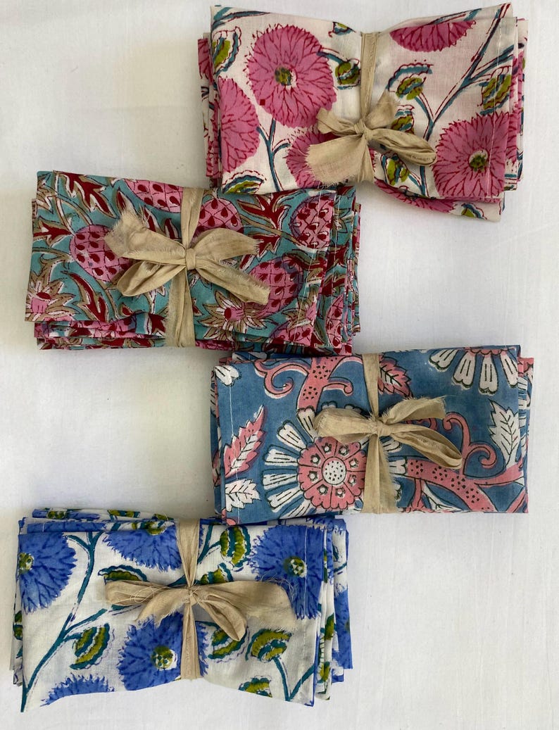 May include: Bundled sets of floral print napkins tied with tan ribbon. The napkins feature pink, blue, and white floral patterns. The sets are neatly stacked, showcasing the variety of designs.