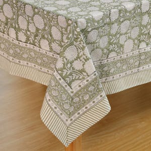 May include: A sage green tablecloth with a white floral design. The border has a striped pattern and floral details. The cloth is draped over a wooden table, highlighting its design and texture.