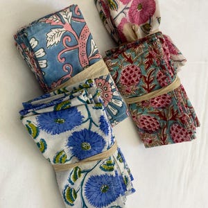 May include: Four bundles of patterned cloth napkins. Each bundle is tied with a beige band. The napkins feature floral and botanical designs in shades of blue, pink, and red on a white background. The napkins are likely made of cotton or linen.