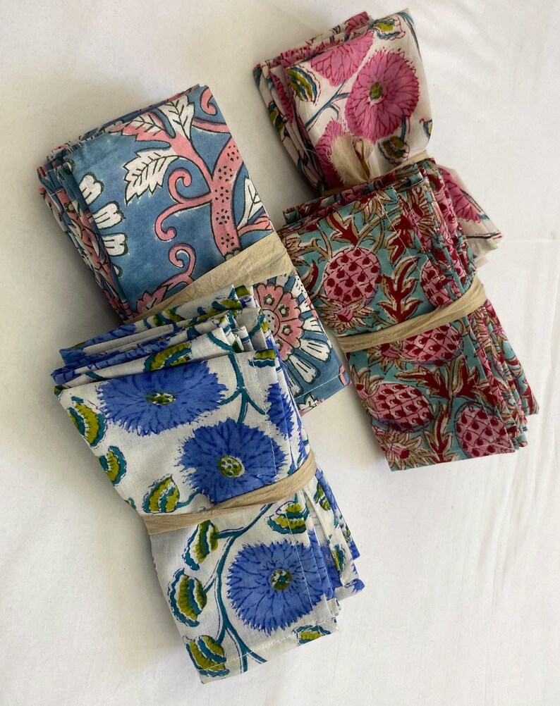 May include: Four bundles of patterned cloth napkins. Each bundle is tied with a beige band. The napkins feature floral and botanical designs in shades of blue, pink, and red on a white background. The napkins are likely made of cotton or linen.