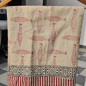 May include: Beige fabric with a repeating pattern of red fish and vertical shapes. The bottom border features a geometric design in black and red stripes. The fabric is hanging on a wooden hanger.