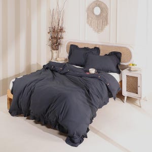 Könnte beinhalten: Ein dunkelblaues Bettwäsche-Set mit gerüschter Bettdecke und passenden Kissenbezügen. Die Bettwäsche befindet sich auf einem Holzrahmen mit einem gewebten Kopfteil. Ein weißer Nachttisch mit einer gewebten Tür steht neben dem Bett.