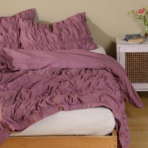 May include: A bed with a mauve-colored comforter and matching pillowcases. The bedding has a ruffled design and wooden buttons. The bed is made up with a white fitted sheet and sits on a wooden frame.