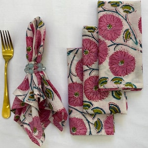 Op de afbeelding: Een set servetten met bloemenprint, een gouden vork en een servetring. De servetten hebben een herhalend patroon van roze bloemen en groene bladeren op een cr&egrave;mekleurige achtergrond. De servetring is lichtblauw.