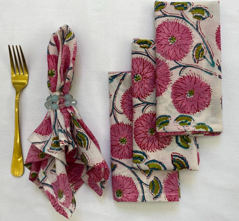Op de afbeelding: Een set servetten met bloemenprint, een gouden vork en een servetring. De servetten hebben een herhalend patroon van roze bloemen en groene bladeren op een cr&egrave;mekleurige achtergrond. De servetring is lichtblauw.
