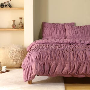 May include: A bed with a mauve ruched duvet cover and matching pillowcases. The bedding is made of a soft fabric and has a textured appearance. The bed is set against a neutral-colored wall and a white rug.