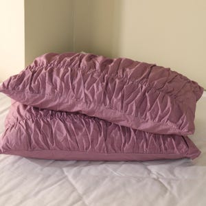 May include: Two mauve-colored decorative pillows stacked on a white bed. The pillows have a ruched, textured design across the top. The pillows are rectangular and appear to be made of a soft fabric.